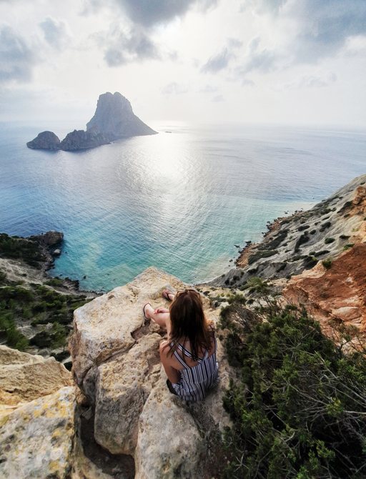 Amazing seascape, girl sitting on the rock and looking at the magic rock Es Vedra Island at sunset, Ibiza, Balearic Island, Spain