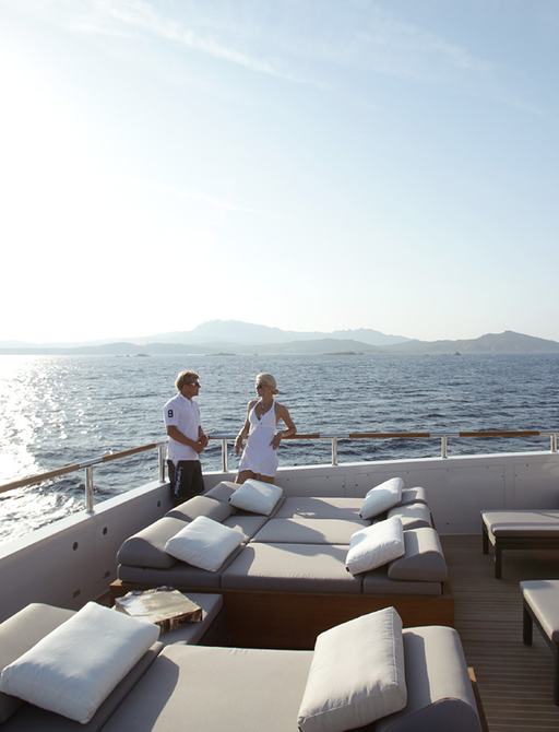 Couple stood on deck of superyacht GEMS II talking as sunsets with sunloungers in foreground
